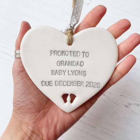 Pearlised white clay hanging heart with baby feet cut out of the bottom and grey personalisation, the heart is personalised with PROMOTED TO GRANDAD BABY LYONS DUE DECEMBER 2025
