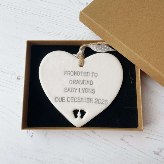 Pearlised white clay hanging heart with baby feet cut out of the bottom and grey personalisation, the heart is personalised with PROMOTED TO GRANDAD BABY LYONS DUE DECEMBER 2025