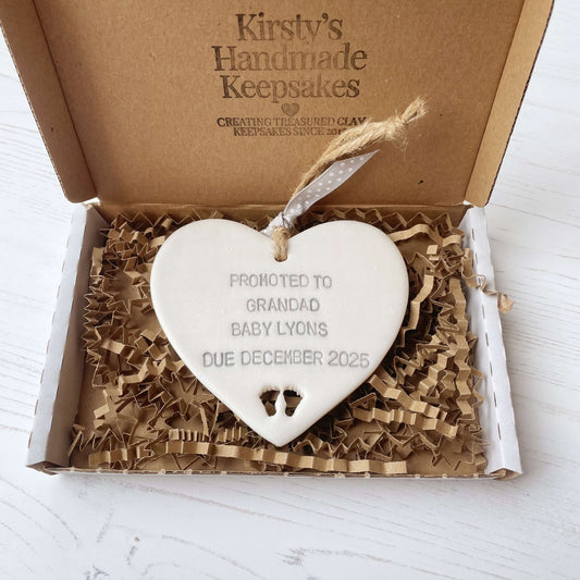 Pearlised white clay hanging heart with baby feet cut out of the bottom and grey personalisation, the heart is personalised with PROMOTED TO GRANDAD BABY LYONS DUE DECEMBER 2025