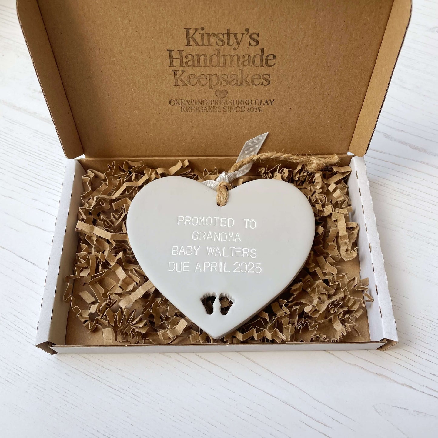 Grey clay hanging heart with baby feet cut out of the bottom and white personalisation, the heart is personalised with PROMOTED TO GRANDMA BABY WALTERS DUE APRIL 2025