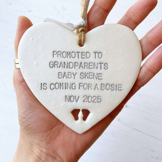 Pearlised white clay hanging heart with baby feet cut out of the bottom and grey personalisation, the heart is personalised with PROMOTED TO GRANDPARENTS BABY SKENE IS COMING FOR A BOSIE NOV 2025