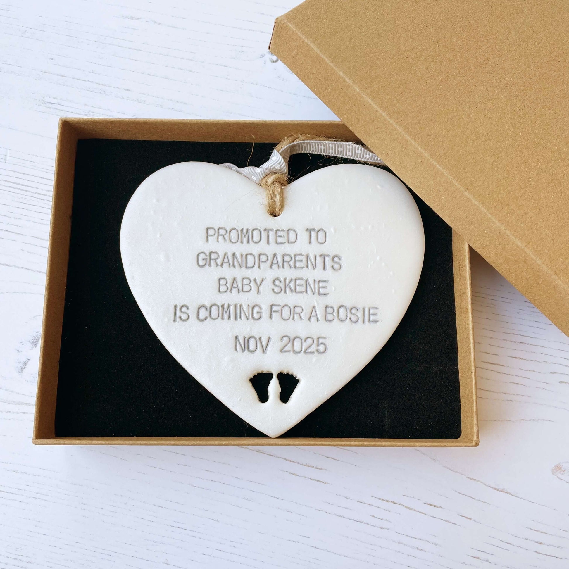 Pearlised white clay hanging heart with baby feet cut out of the bottom and grey personalisation, the heart is personalised with PROMOTED TO GRANDPARENTS BABY SKENE IS COMING FOR A BOSIE NOV 2025
