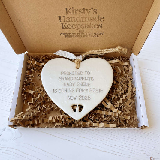 Pearlised white clay hanging heart with baby feet cut out of the bottom and grey personalisation, the heart is personalised with PROMOTED TO GRANDPARENTS BABY SKENE IS COMING FOR A BOSIE NOV 2025