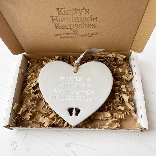 Grey clay hanging heart with baby feet cut out of the bottom and grey personalisation, the heart is personalised with PROMOTED TO GRANDPARENTS BABY TRELEAVEN DUE OCTOBER 2024 In a postal box with brown Kraft shredded zigzag paper