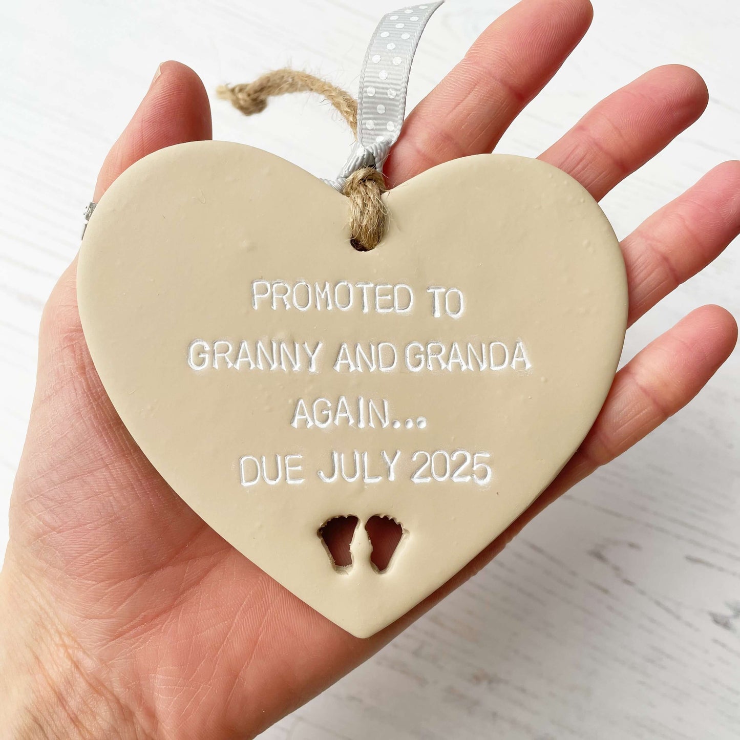 Beige clay hanging heart with baby feet cut out of the bottom and white personalisation, the heart is personalised with PROMOTED TO GRANNY AND GRANDA AGAIN… DUE JULY 2025