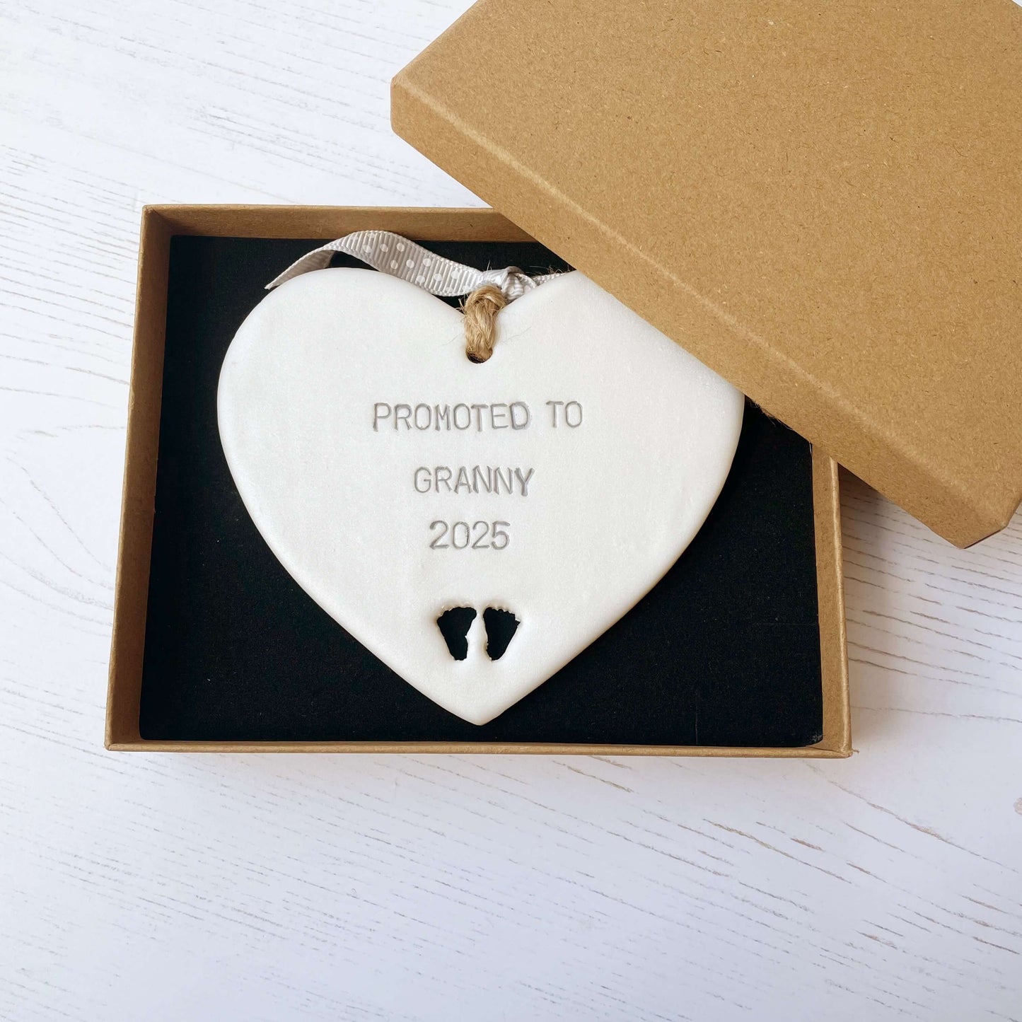 Pearlised white clay hanging heart with baby feet cut out of the bottom and grey personalisation, the heart is personalised with PROMOTED TO GRANNY 2025