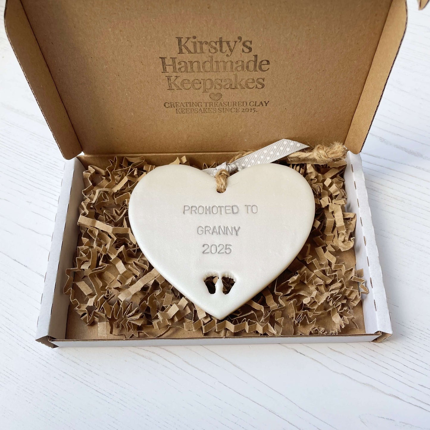 Pearlised white clay hanging heart with baby feet cut out of the bottom and grey personalisation, the heart is personalised with PROMOTED TO GRANNY 2025