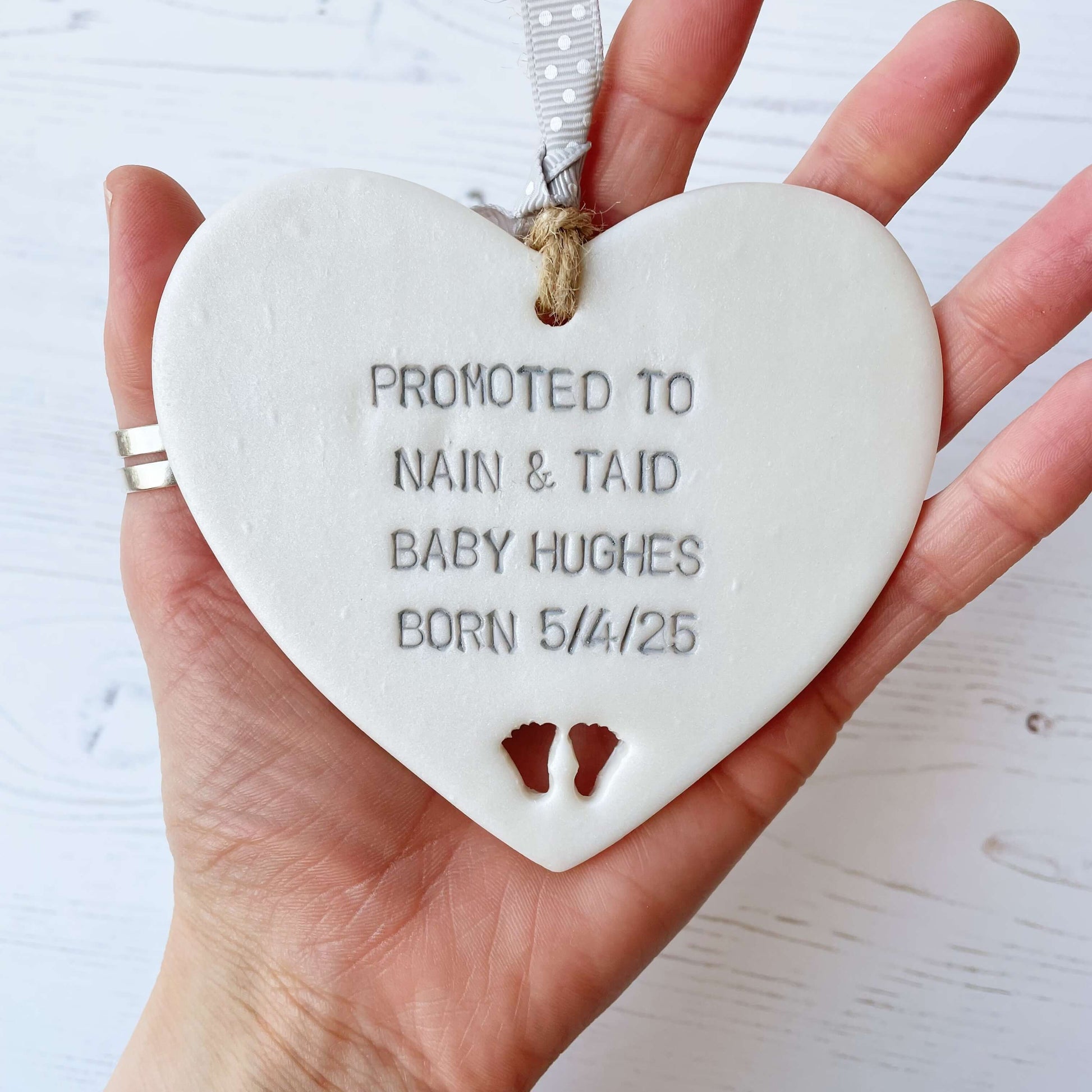 Pearlised white clay hanging heart with baby feet cut out of the bottom and grey personalisation, the heart is personalised with PROMOTED TO NAIN & TAID BABY HUGHES BORN 5/4/25