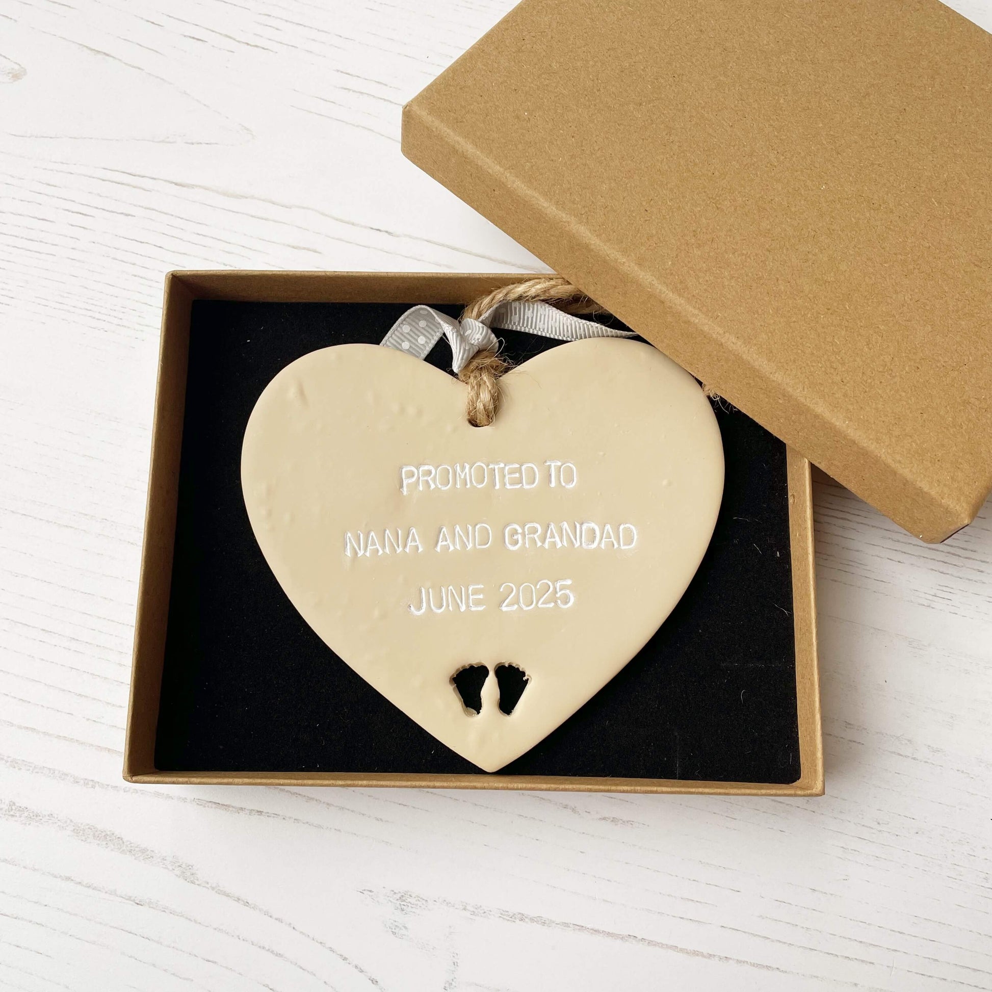Beige clay hanging heart with baby feet cut out of the bottom and white personalisation, the heart is personalised with PROMOTED TO NANA AND GRANDAD JUNE 2025