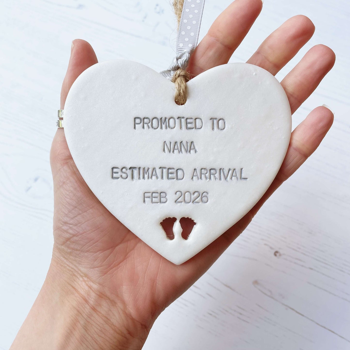 Pearlised white clay hanging heart with baby feet cut out of the bottom and grey personalisation, the heart is personalised with PROMOTED TO NANA ESTIMATED ARRIVAL FEB 2026