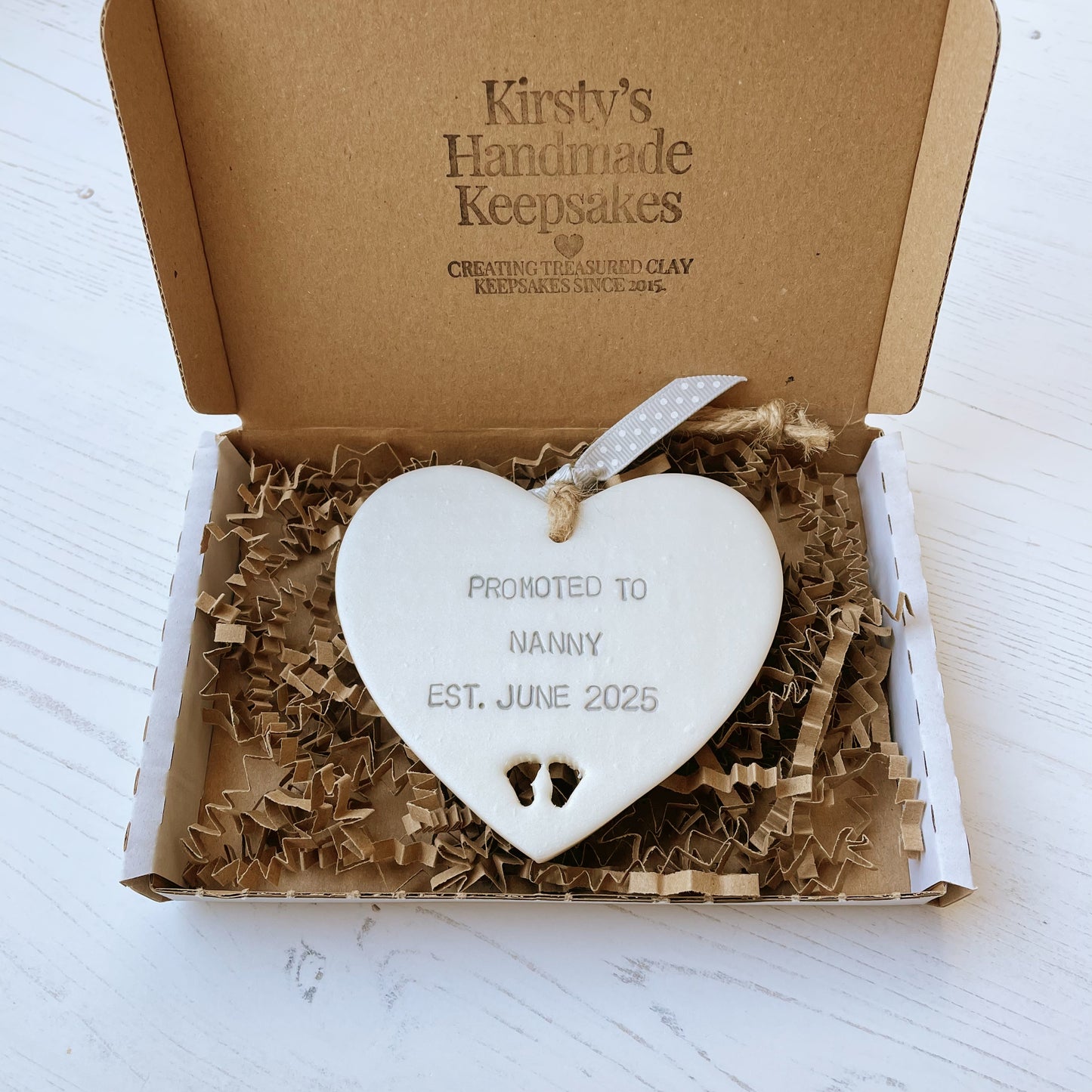 Pearlised white clay hanging heart with baby feet cut out of the bottom and grey personalisation, the heart is personalised with PROMOTED TO NANNY EST. JUNE 2025