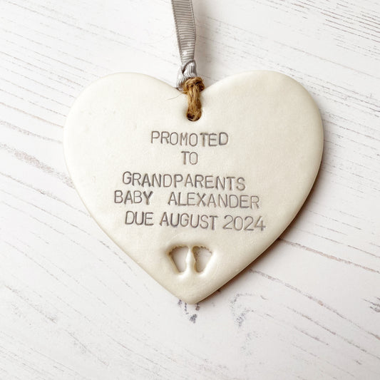 Pearlised white clay hanging heart with baby feet cut out of the bottom and grey personalisation, the heart is personalised with PROMOTED TO GRANDPARENTS BABY ALEXANDER DUE AUGUST 2024