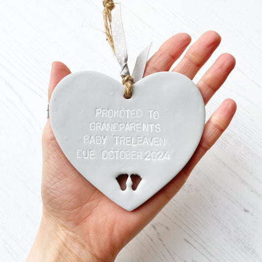 Grey clay hanging heart with baby feet cut out of the bottom and grey personalisation, the heart is personalised with PROMOTED TO GRANDPARENTS BABY TRELEAVEN DUE OCTOBER 2024