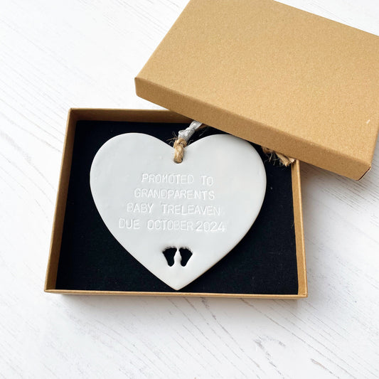 Grey clay hanging heart with baby feet cut out of the bottom and grey personalisation, the heart is personalised with PROMOTED TO GRANDPARENTS BABY TRELEAVEN DUE OCTOBER 2024 In a brown Kraft luxury gift box