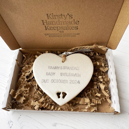 Pearlised white clay hanging heart with baby feet cut out of the bottom and grey personalisation, the heart is personalised with NANNY & GRANDAD BABY TRELEAVEN DUE OCTOBER 2024 In a postal box with brown Kraft shredded zigzag paper