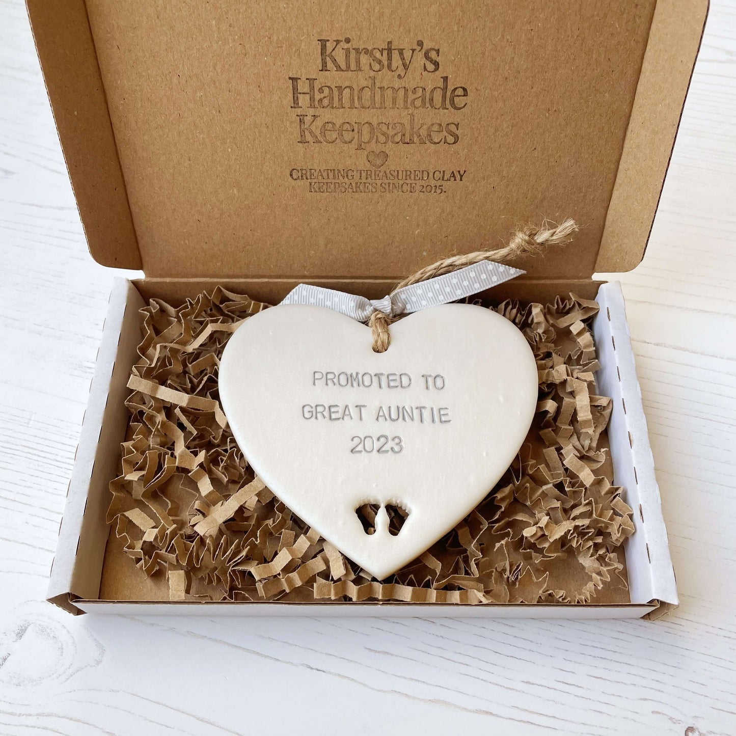 Pearlised white clay hanging heart with baby feet cut out of the bottom and grey personalisation, the heart is personalised with PROMOTED TO GREAT AUNTIE 2023