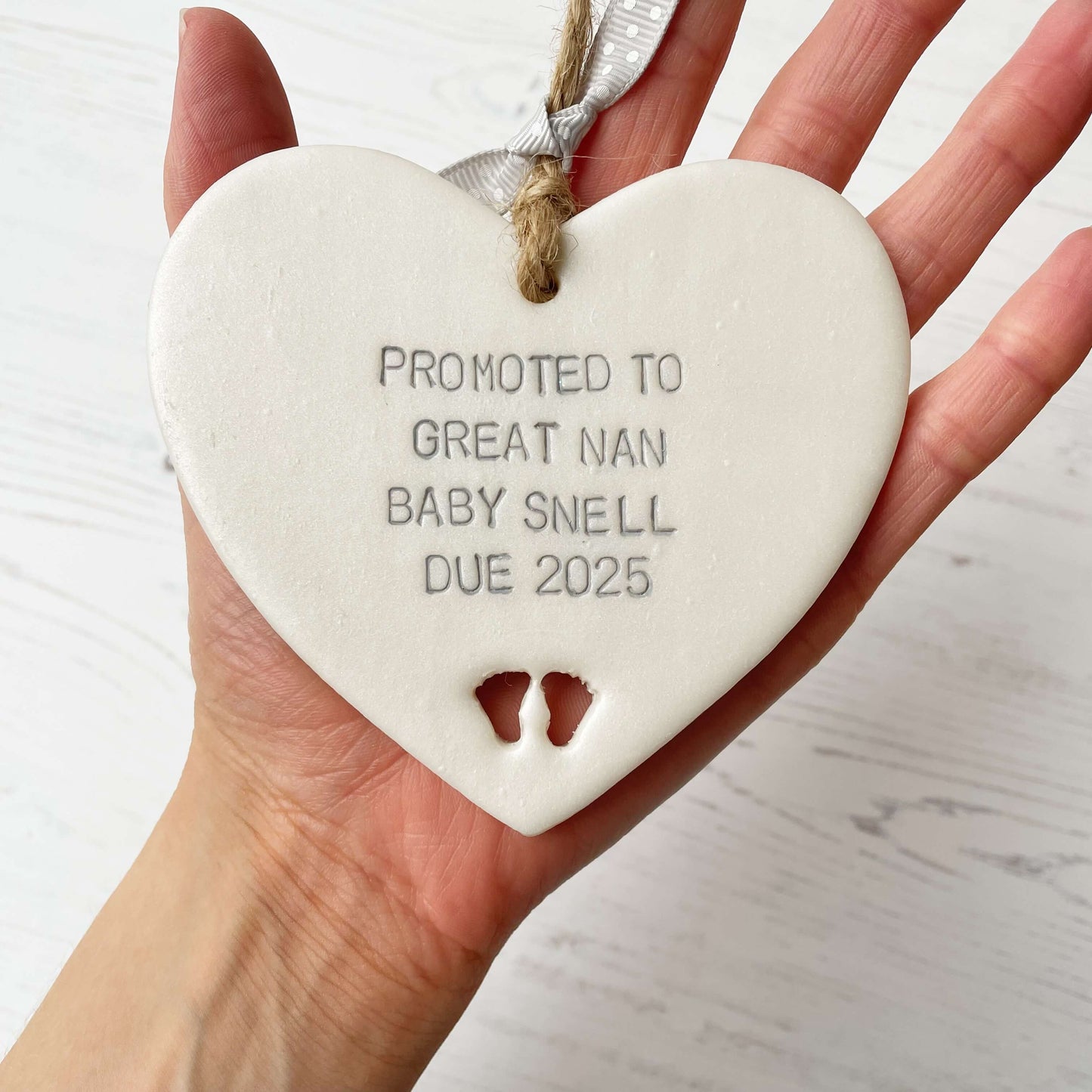 Pearlised white clay hanging heart with baby feet cut out of the bottom and grey personalisation, the heart is personalised with PROMOTED TO GREAT NAN BABY SNELL DUE 2025