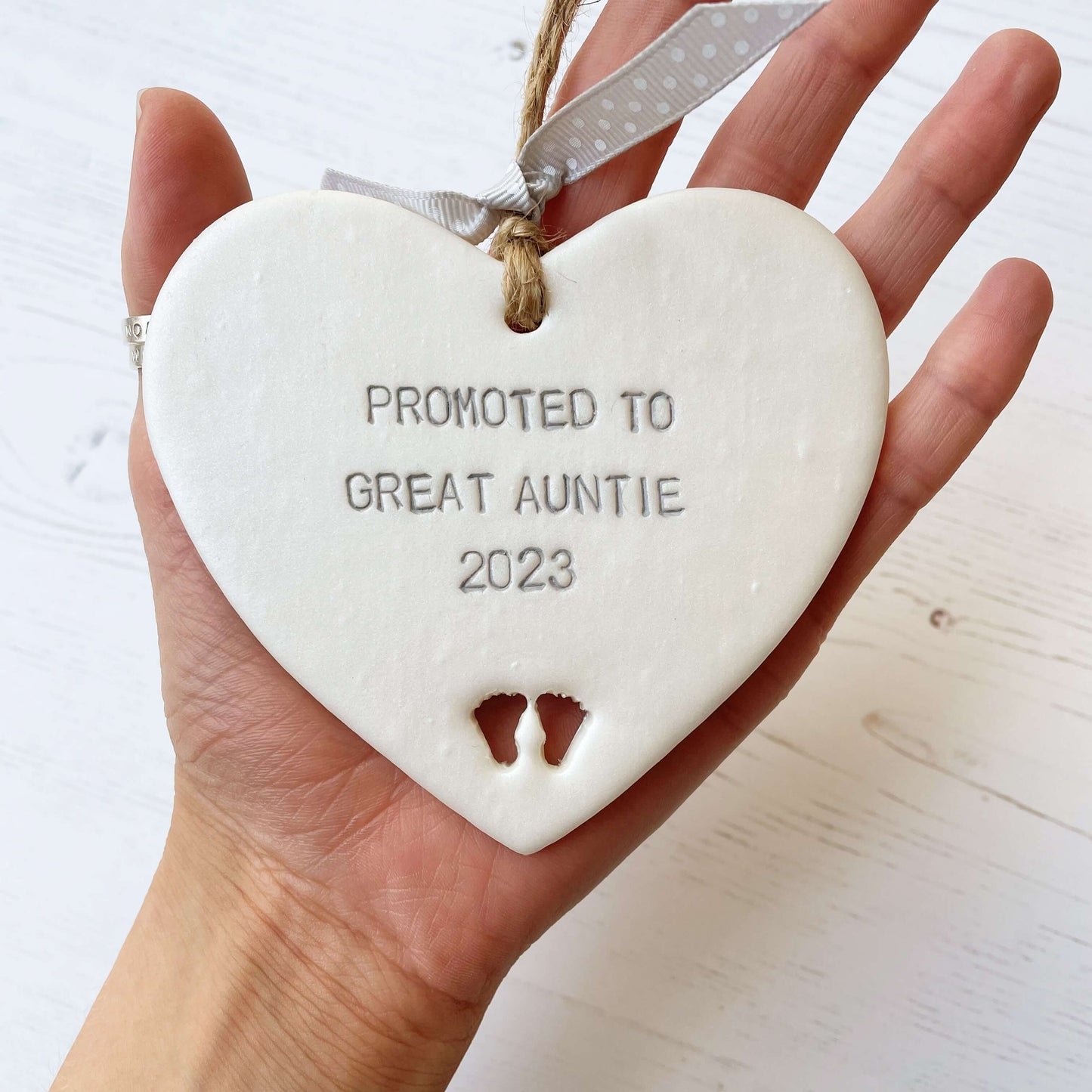Pearlised white clay hanging heart with baby feet cut out of the bottom and grey personalisation, the heart is personalised with PROMOTED TO GREAT AUNTIE 2023