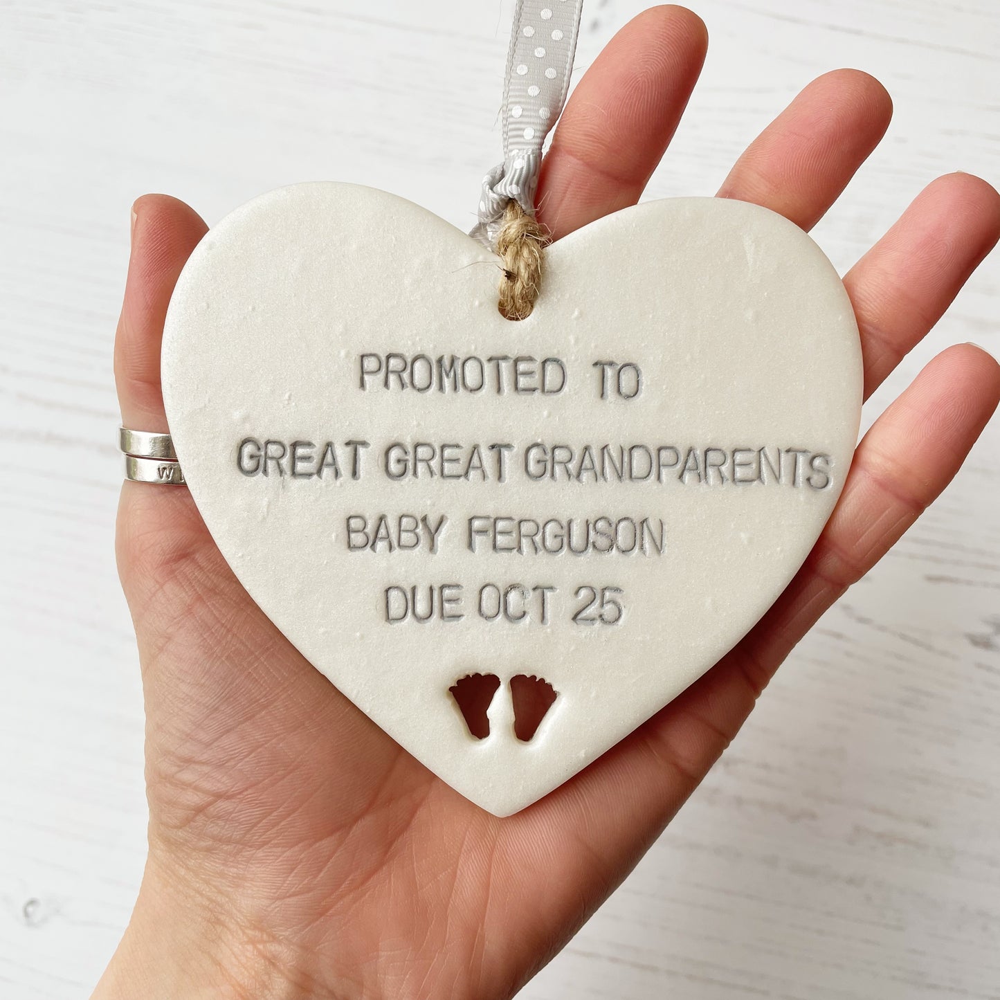 Pearlised white clay hanging heart with baby feet cut out of the bottom and grey personalisation, the heart is personalised with PROMOTED TO GREAT GREAT GRANDPARENTS BABY FERGUSON DUE OCT 2025