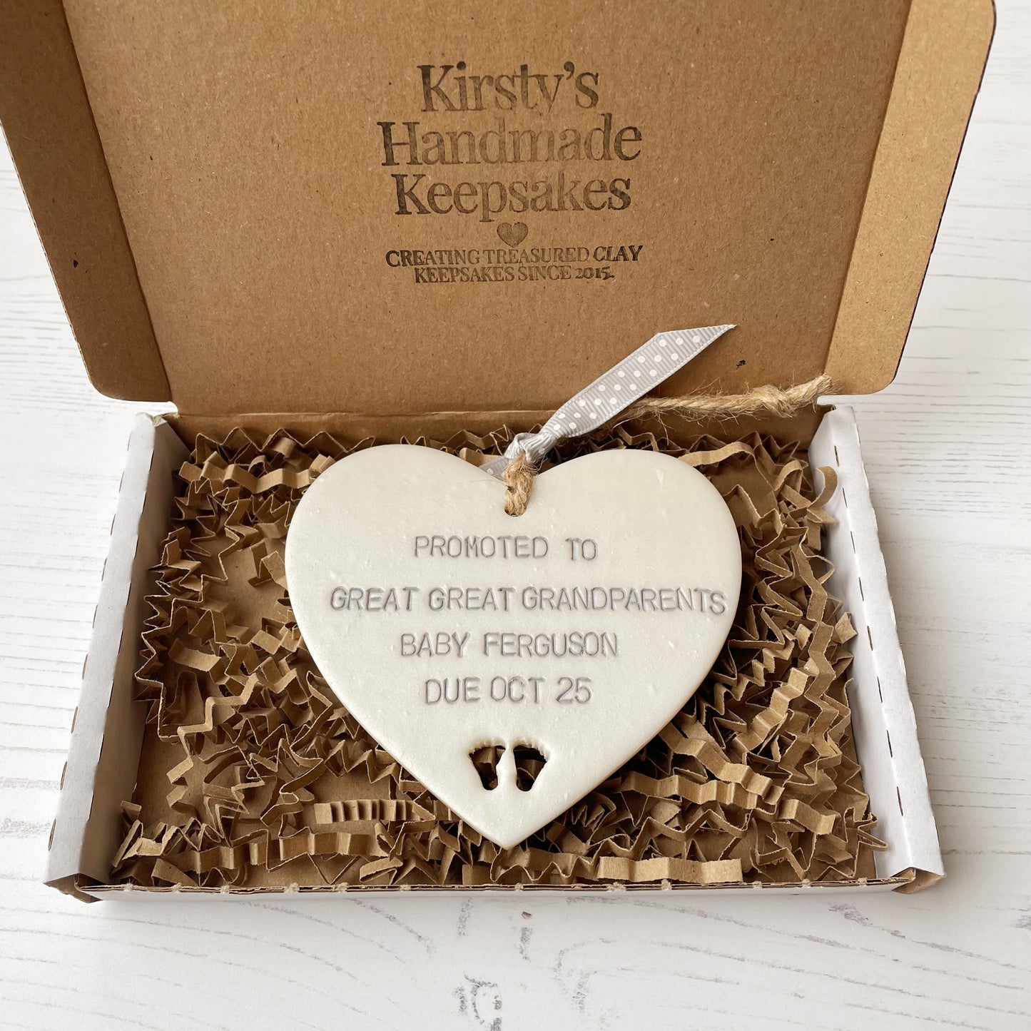 Pearlised white clay hanging heart with baby feet cut out of the bottom and grey personalisation, the heart is personalised with PROMOTED TO GREAT GREAT GRANDPARENTS BABY FERGUSON DUE OCT 2025