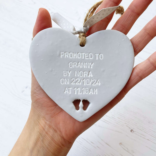 Grey clay hanging heart with baby feet cut out of the bottom and white personalisation, the heart is personalised with PROMOTED TO GRANNY BY NORA ON 22/10/24 AT 11.16 AM