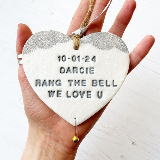 Personalised cancer survivor gift, pearlised white clay hanging heart with a grey lace edge at the top of the heart and a gold bell hanging below, the heart is personalised with 10-01-24 DARCIE RANG THE BELL WE LOVE U