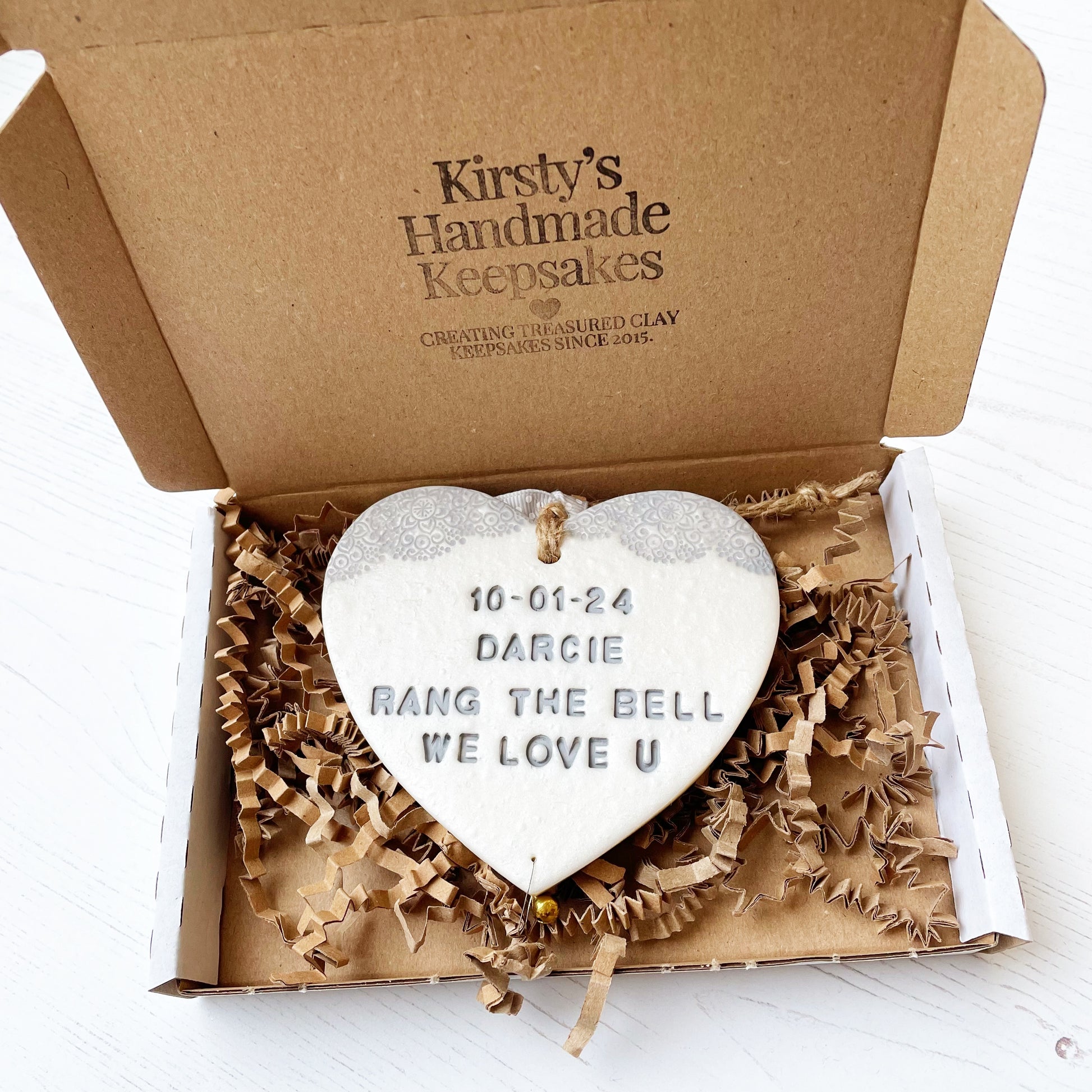 Personalised cancer survivor gift, pearlised white clay hanging heart with a grey lace edge at the top of the heart and a gold bell hanging below, the heart is personalised with 10-01-24 DARCIE RANG THE BELL WE LOVE U. In a white postal box with brown zigzag shredded paper