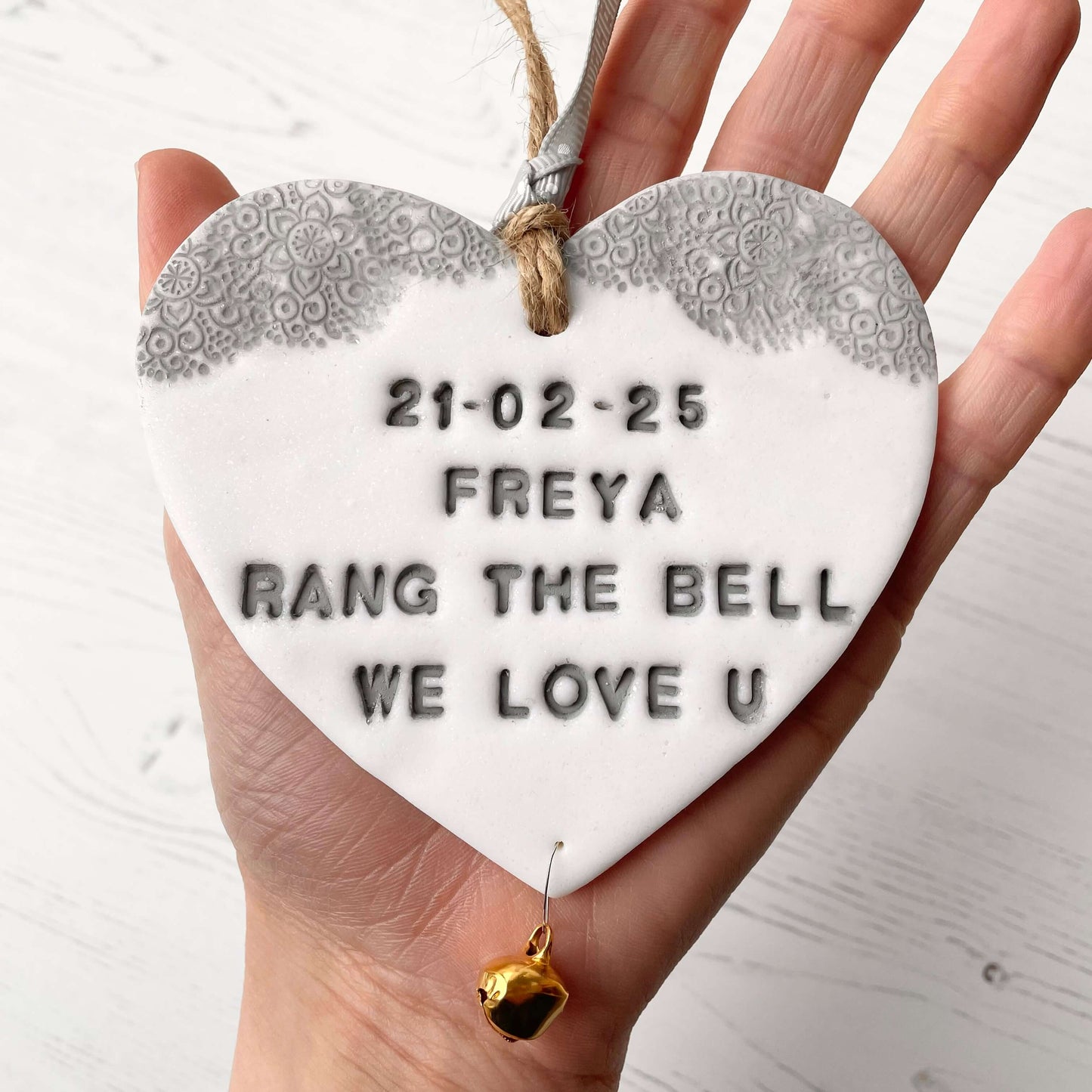 Personalised cancer survivor gift, glitter white clay hanging heart with a grey lace edge at the top of the heart and a gold bell hanging below, the heart is personalised with 21-02-25 FREYA RANG THE BELL WE LOVE U
