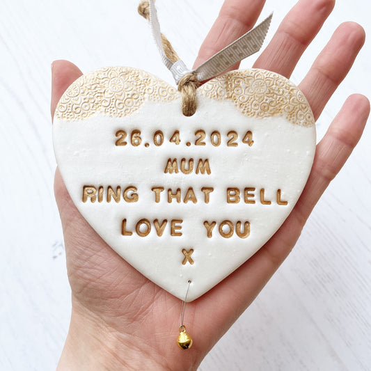 Personalised cancer survivor gift, pearlised white clay hanging heart with a gold lace edge at the top of the heart and a gold bell hanging below, the heart is personalised with 26.04.2024 MUM RING THAT BELL LOVE YOU X