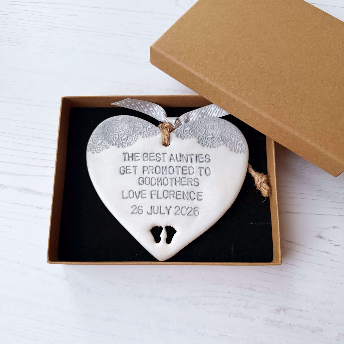 Personalised Godmother gift, pearlised white clay hanging heart with baby feet cut out of the bottom and grey lace edge at the top of the heart, the heart is personalised with THE BEST AUNTIES GET PROMOTED TO GODMOTHERS LOVE FLORENCE 26 JULY 2026
