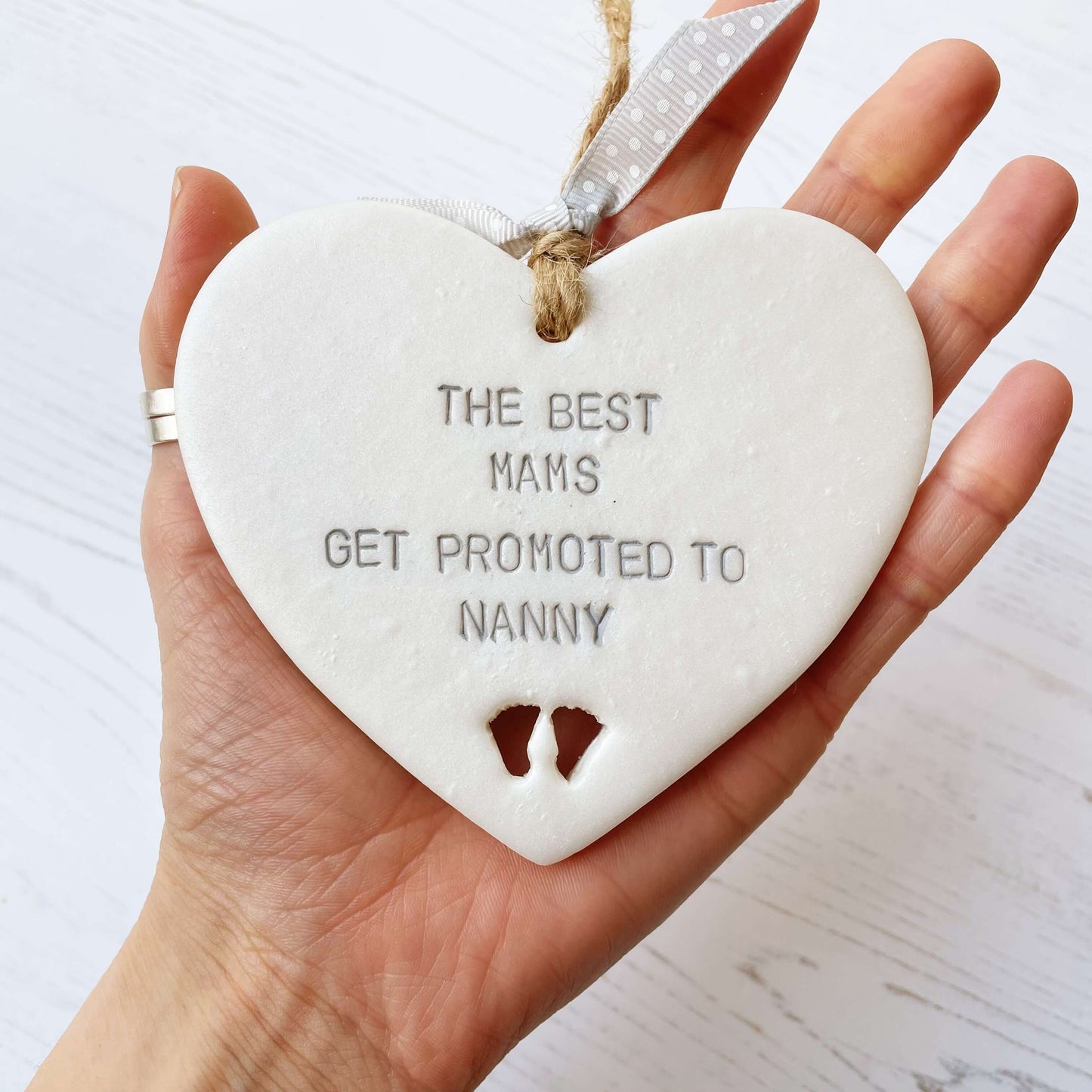 Pearlised white clay hanging heart with baby feet cut out of the bottom and grey personalisation, the heart is personalised with THE BEST MAMS GET PROMOTED TO NANNY
