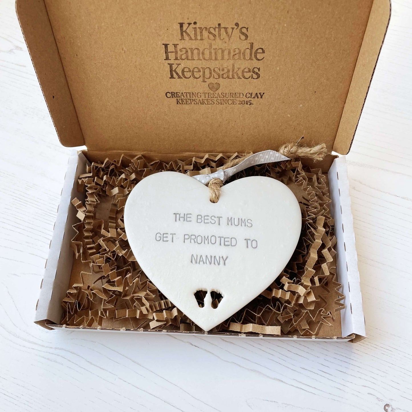 Pearlised white clay hanging heart with baby feet cut out of the bottom and grey personalisation, the heart is personalised with THE BEST MUMS GET PROMOTED TO NANNY