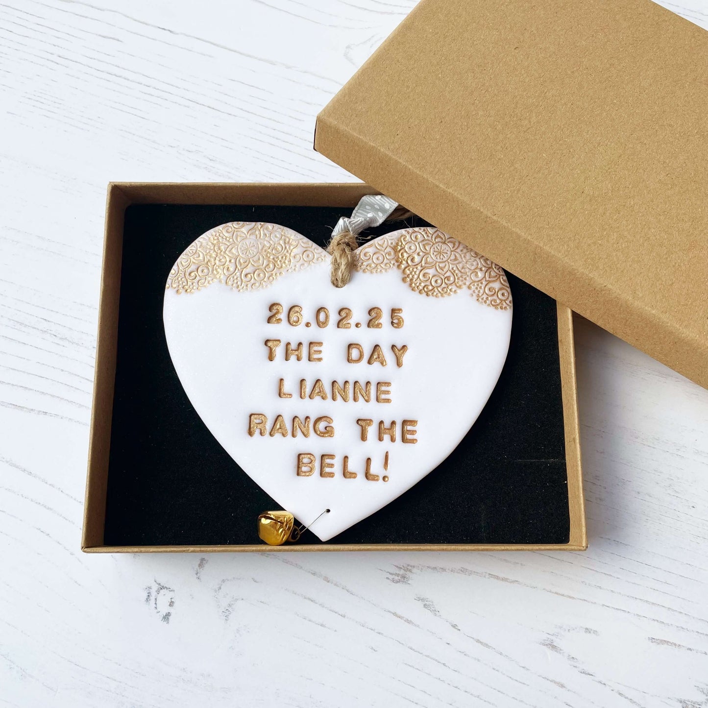 Personalised cancer survivor gift, glitter white clay hanging heart with a gold lace edge at the top of the heart and a gold bell hanging below, the heart is personalised with 26.02.25 THE DAY LIANNE RANG THE BELL!
