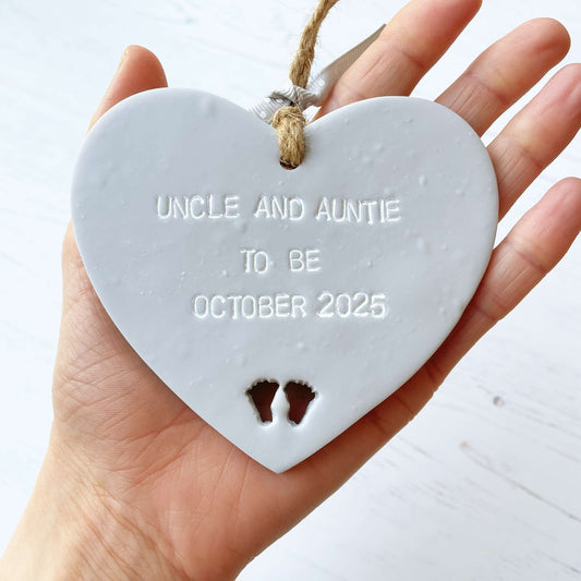 Grey clay hanging heart with baby feet cut out of the bottom and white personalisation, the heart is personalised with UNCLE AND AUNTIE TO BE OCTOBER 2025
