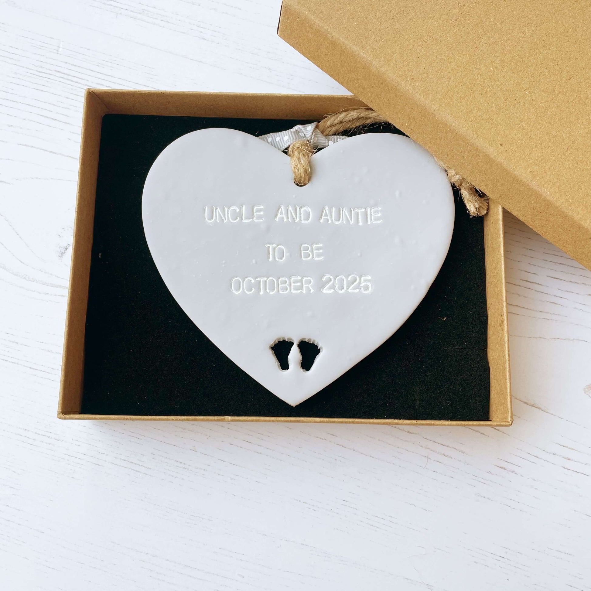 Grey clay hanging heart with baby feet cut out of the bottom and white personalisation, the heart is personalised with UNCLE AND AUNTIE TO BE OCTOBER 2025