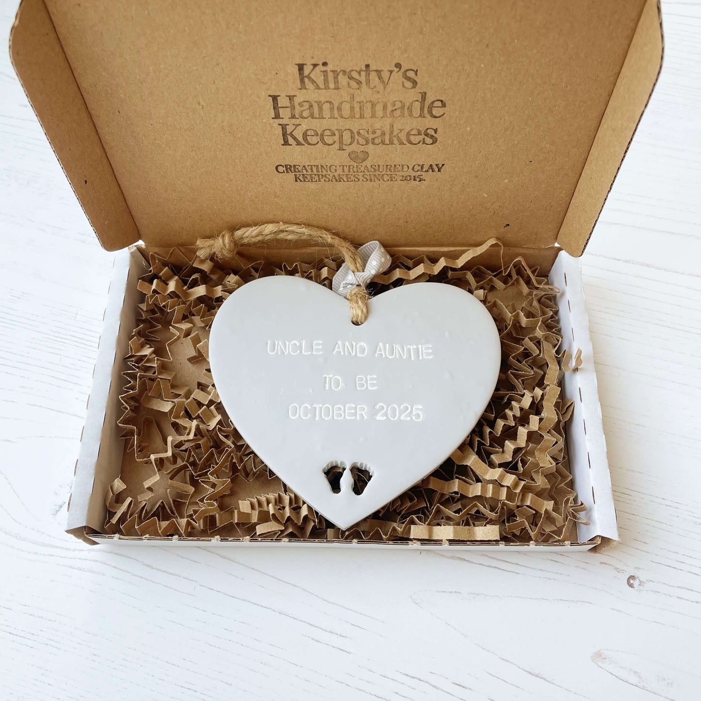 Grey clay hanging heart with baby feet cut out of the bottom and white personalisation, the heart is personalised with UNCLE AND AUNTIE TO BE OCTOBER 2025