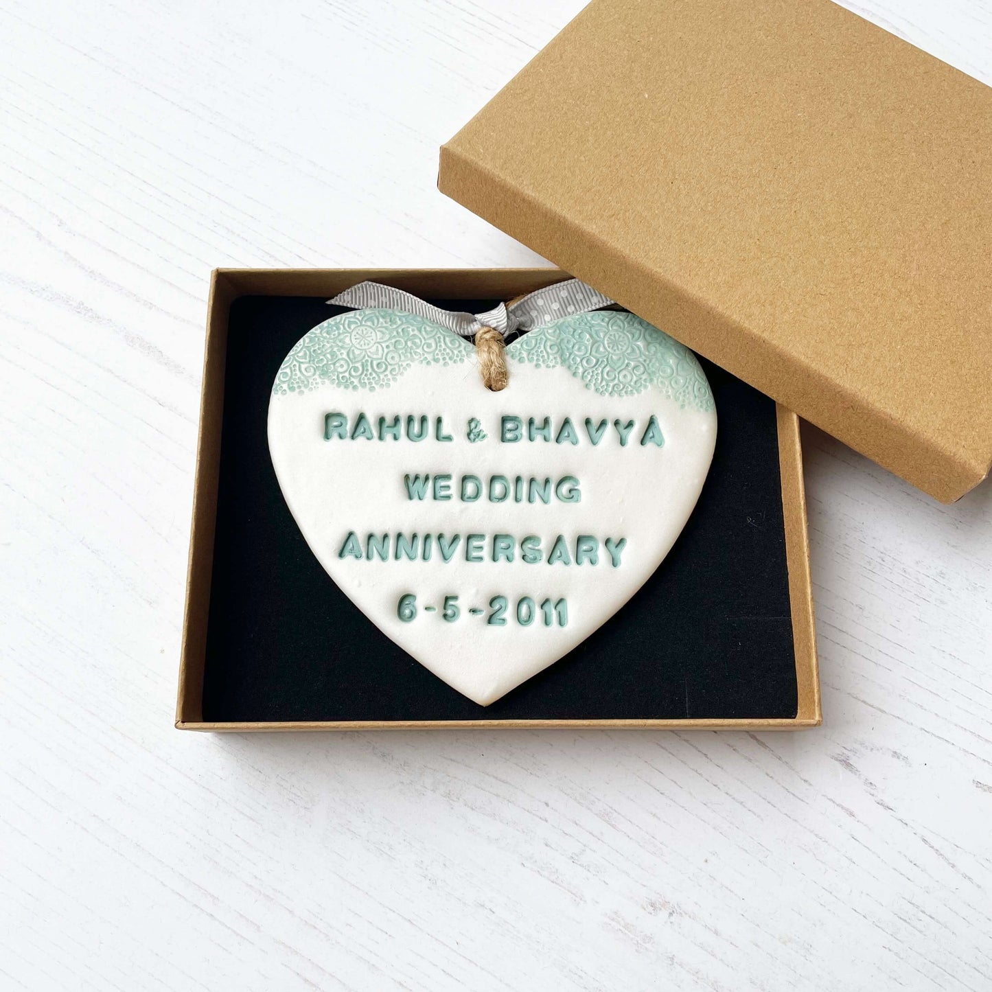 Personalised 13th wedding anniversary gift, pearlised white clay hanging heart with a sage green lace edge at the top of the heart, the heart is personalised with RAHUL & BHAVYA WEDDING ANNIVERSARY 6-5-2011