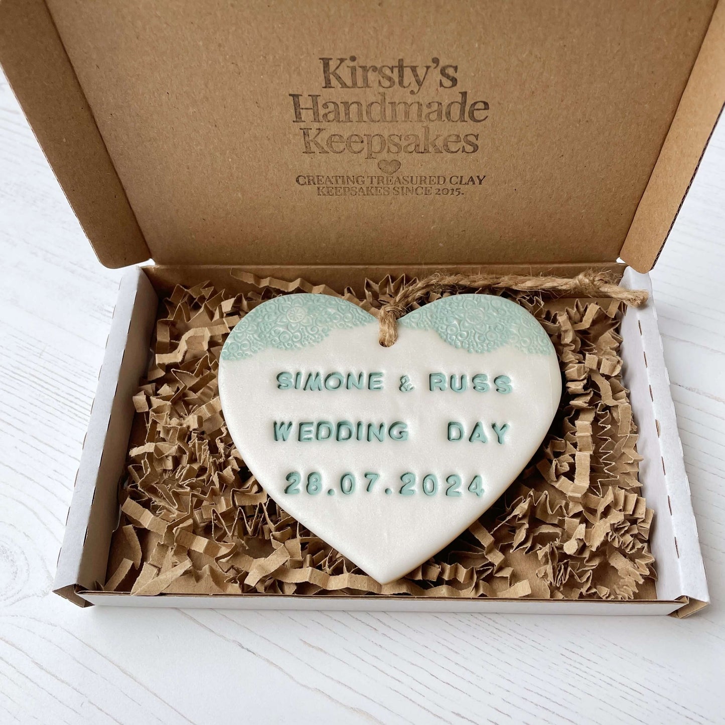 Personalised wedding gift, pearlised white clay heart with a sage green lace edge at the top of the heart with twine to hang, the heart is personalised with SIMONE & RUSS WEDDING DAY 28.07.2024
