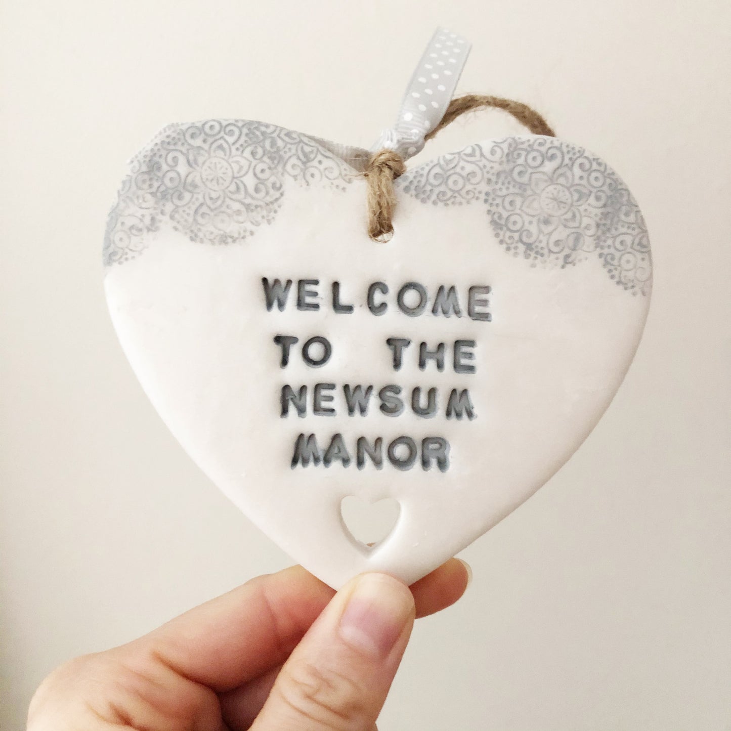 Personalised housewarming new home gift, pearlised white clay heart with a grey lace edge at the top of the heart and a heart cut out at the bottom with jute twine for hanging, the heart is personalised with WELCOME TO THE NEWSUM MANOR