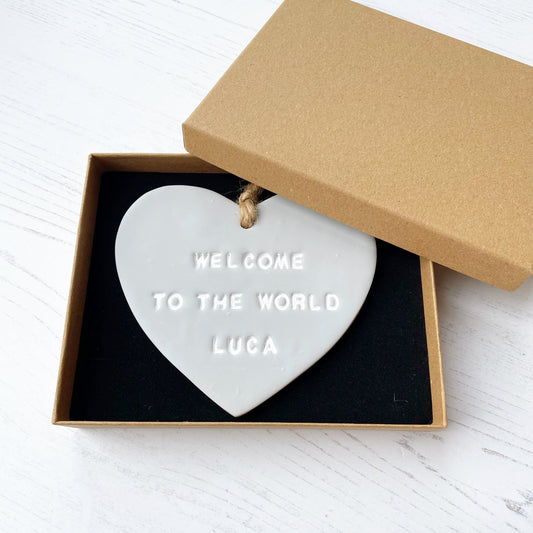Personalised baby announcement heart sign, in grey clay hanging heart with a white writing, the heart is personalised with WELCOME TO THE WORLD LUCA in a brown Kraft luxury gift box