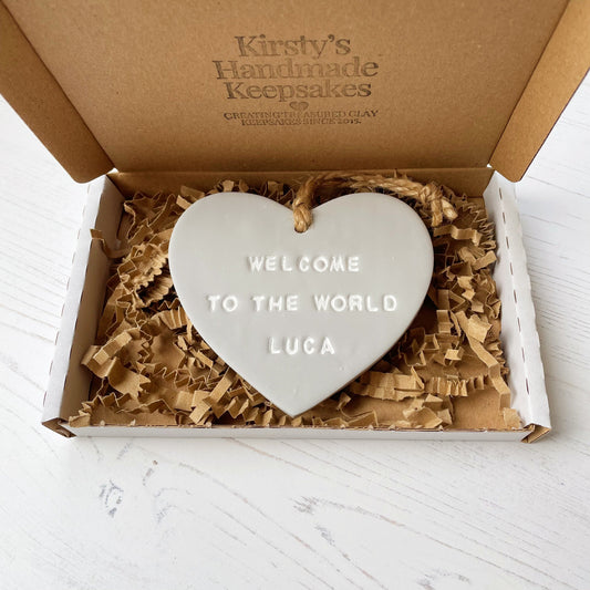 Personalised baby announcement heart sign, in grey clay hanging heart with a white writing, the heart is personalised with WELCOME TO THE WORLD LUCA in a postal box with brown Kraft shredded zigzag paper