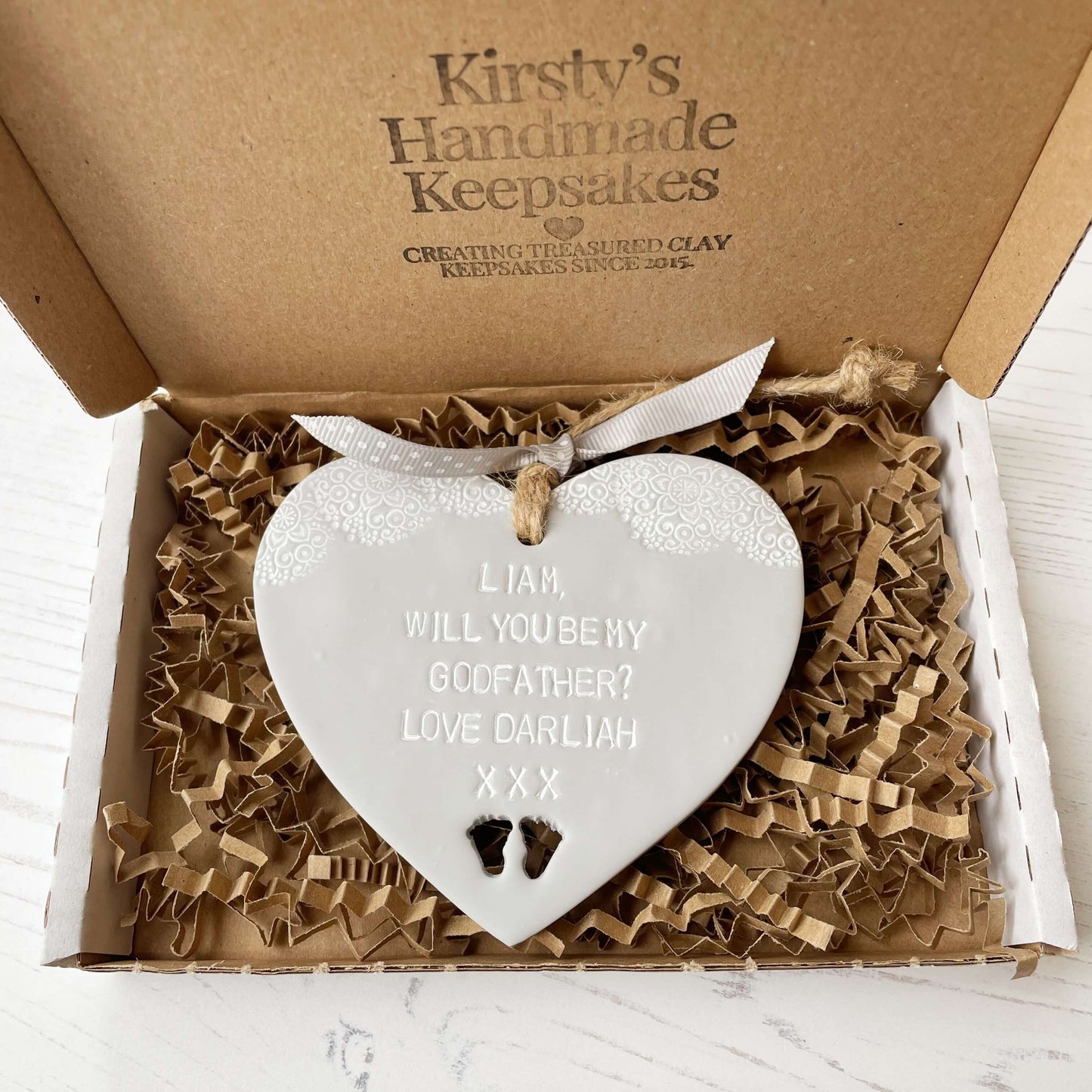 Personalised Godfather proposal gift, grey clay hanging heart with baby feet cut out of the bottom and white lace edge at the top of the heart, the heart is personalised with LIAM, WILL YOU BE MY GODFATHER? LOVE DARLIAH XXX