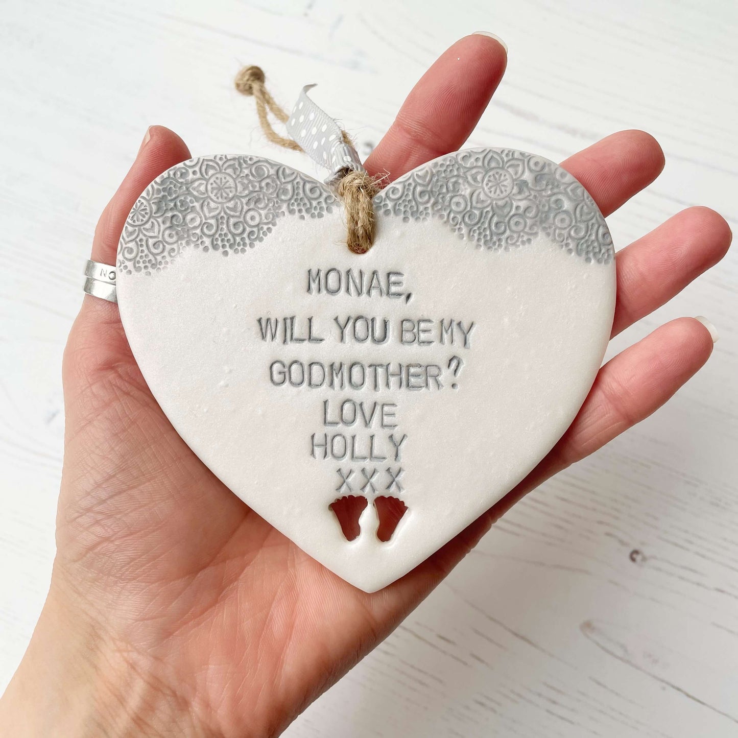 Personalised Godmother gift, pearlised white clay hanging heart with baby feet cut out of the bottom and grey lace edge at the top of the heart, the heart is personalised with MONAE, WILL YOU BE MY GODMOTHER? LOVE HOLLY XXX