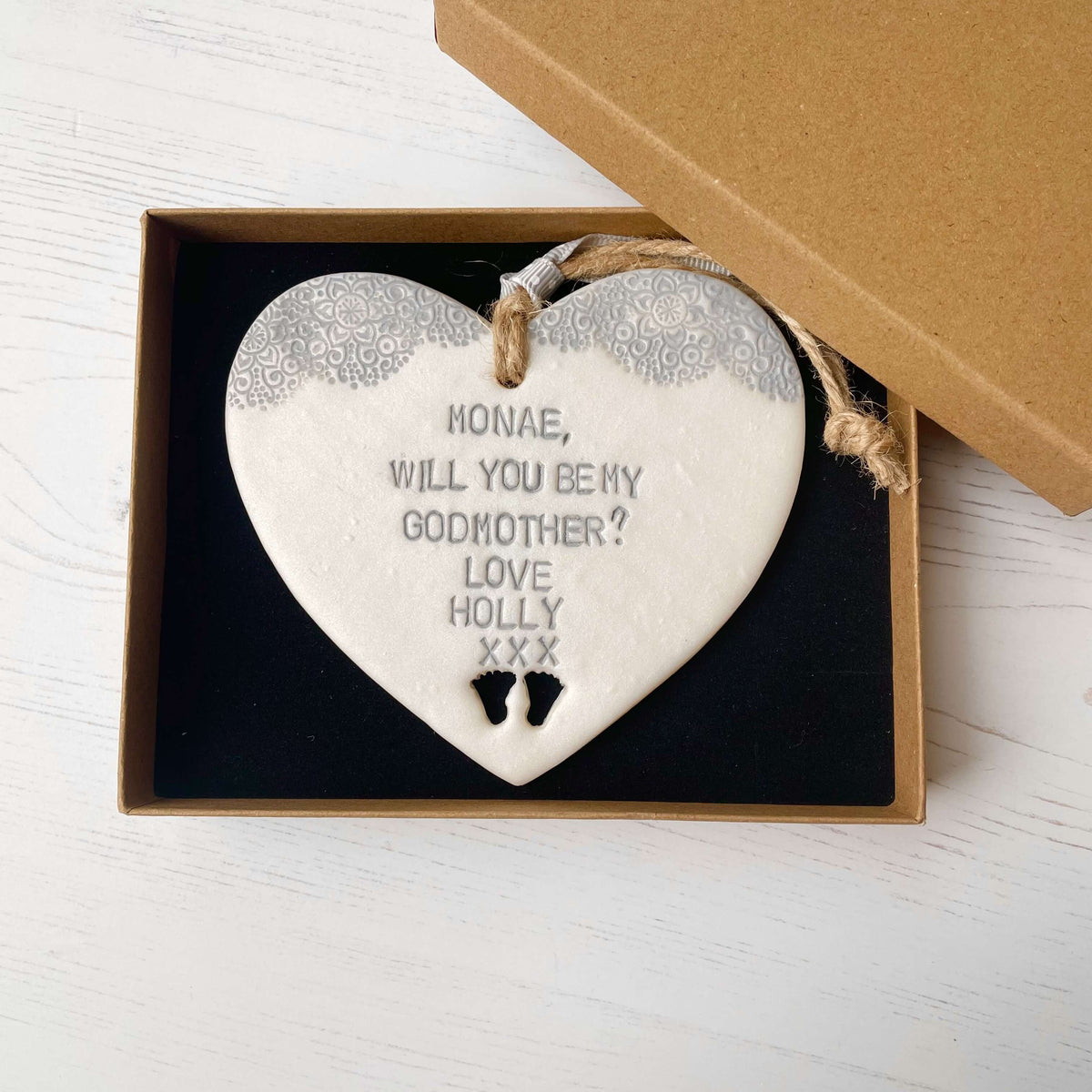 Personalised Godmother gift, pearlised white clay hanging heart with baby feet cut out of the bottom and grey lace edge at the top of the heart, the heart is personalised with MONAE, WILL YOU BE MY GODMOTHER? LOVE HOLLY XXX