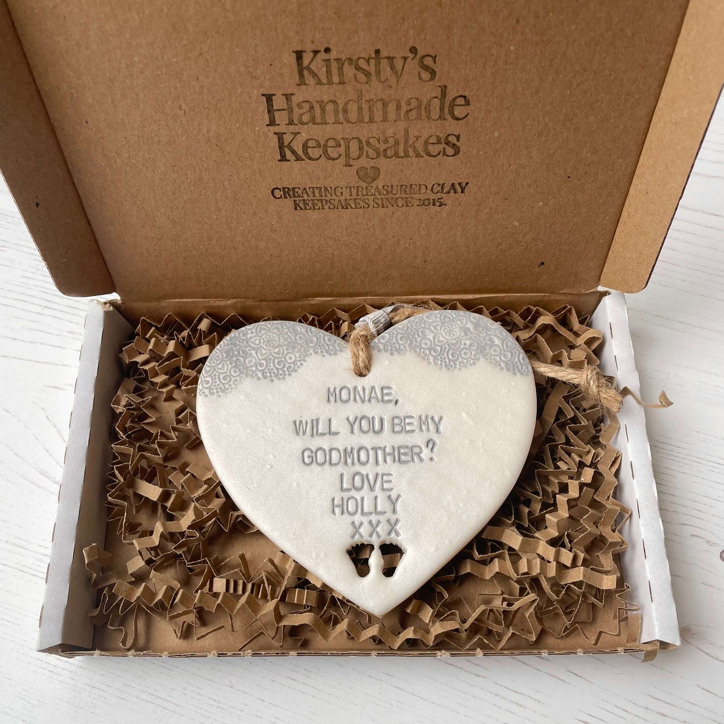 Personalised Godmother gift, pearlised white clay hanging heart with baby feet cut out of the bottom and grey lace edge at the top of the heart, the heart is personalised with MONAE, WILL YOU BE MY GODMOTHER? LOVE HOLLY XXX