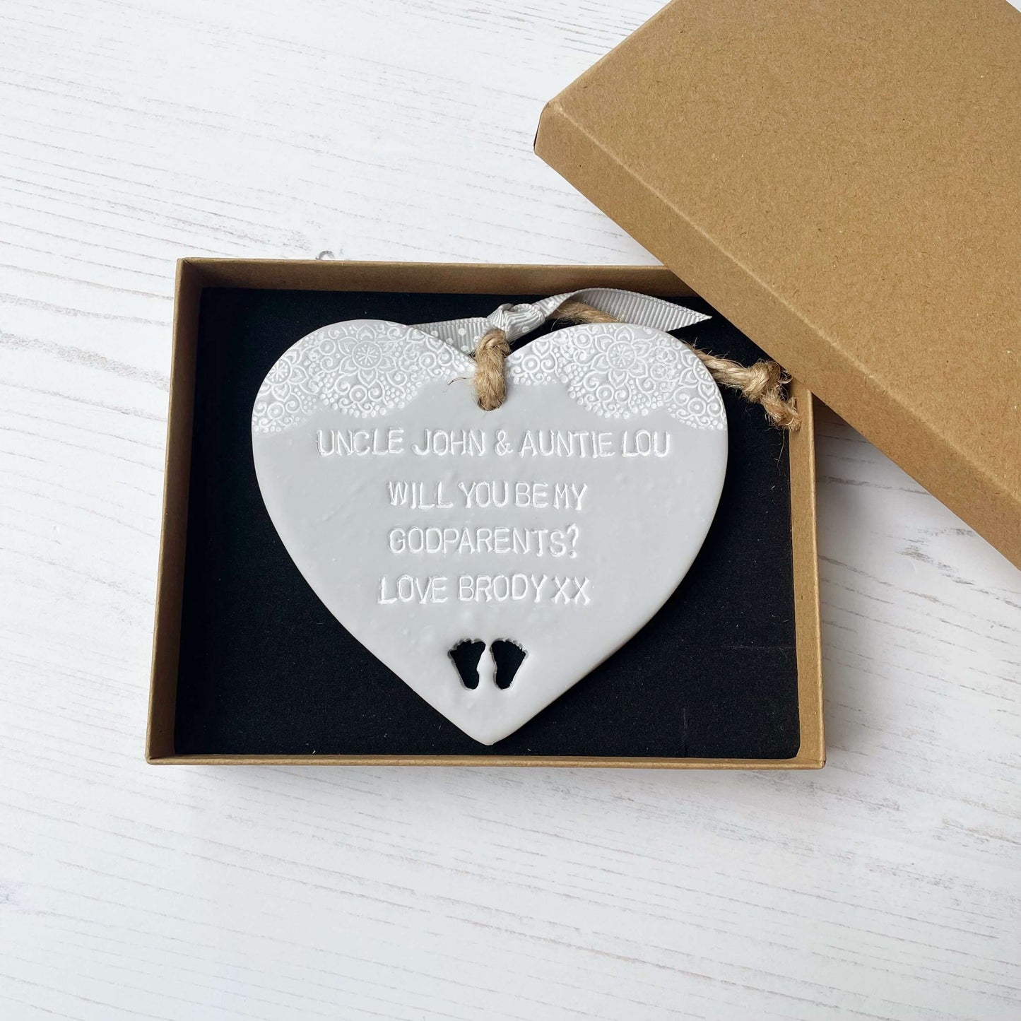 Personalised Godparents proposal gift, grey clay hanging heart with baby feet cut out of the bottom and white lace edge at the top of the heart, the heart is personalised with UNCLE JOHN & AUNTIE LOU WILL YOU BE MY GODPARENTS? LOVE BRODY XX