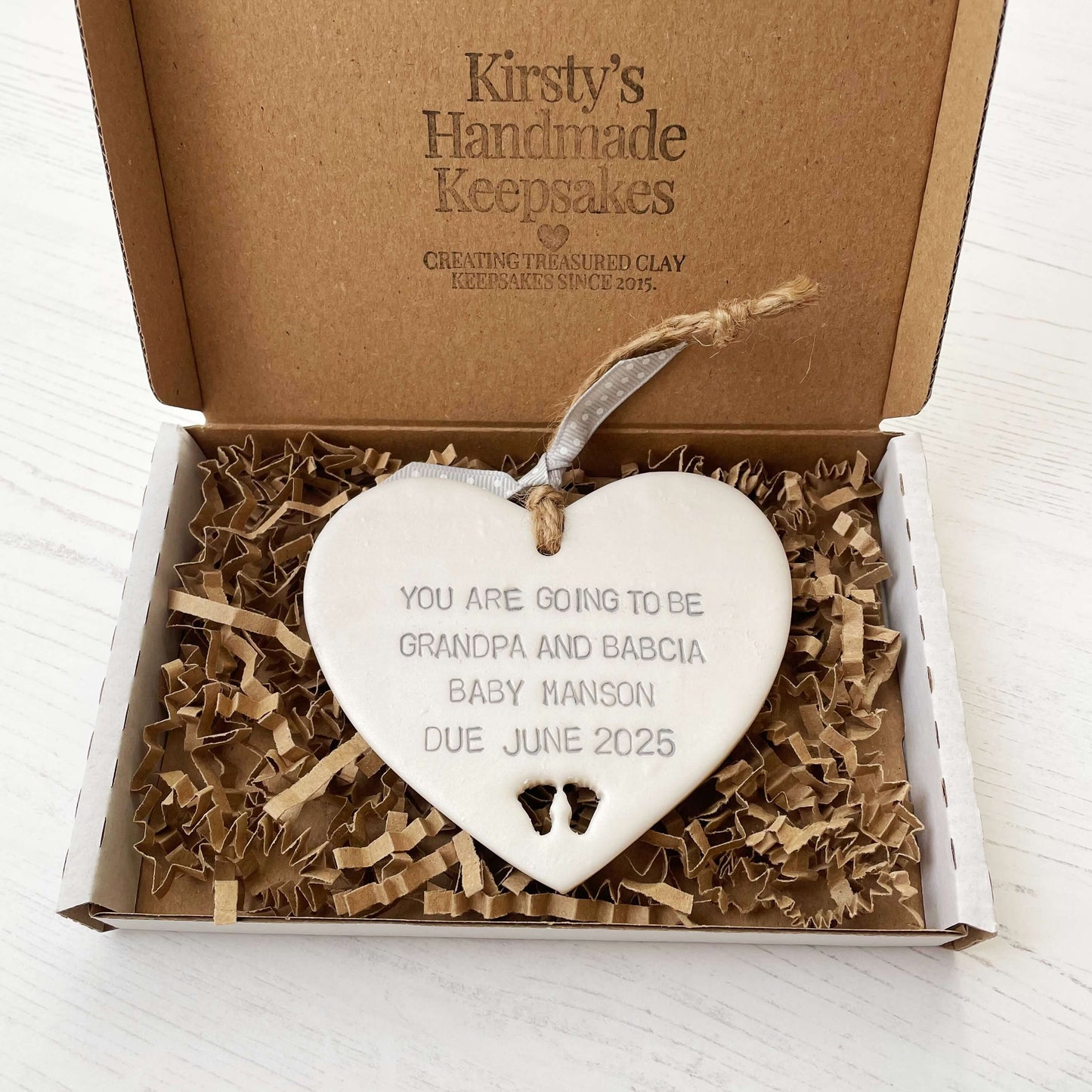 Pearlised white clay hanging heart with baby feet cut out of the bottom and grey personalisation, the heart is personalised with YOU ARE GOING TO BE GRANDPARENTS BABY MANSON DUE JUNE 2025