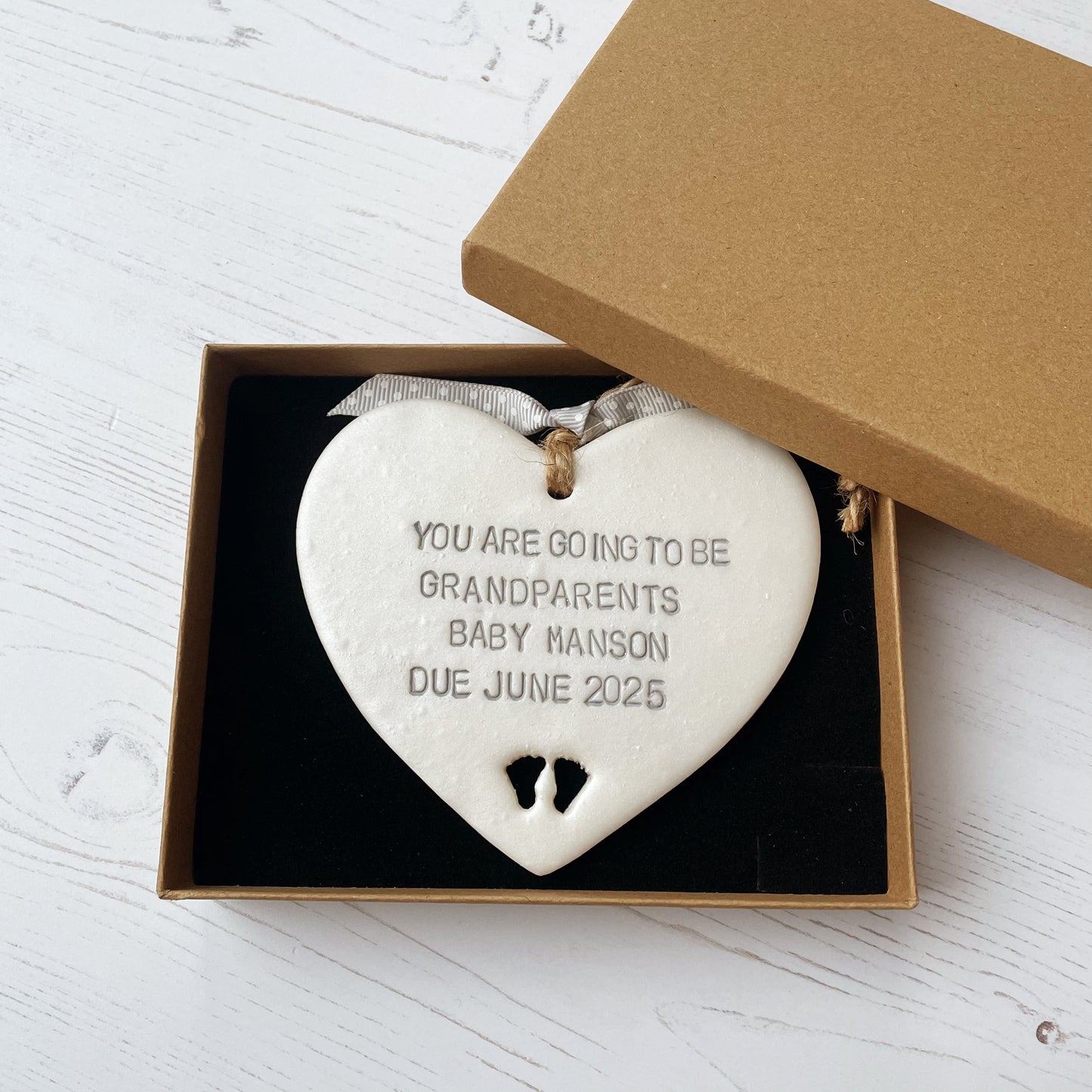 Pearlised white clay hanging heart with baby feet cut out of the bottom and grey personalisation, the heart is personalised with YOU ARE GOING TO BE GRANDPARENTS BABY MANSON DUE JUNE 2025