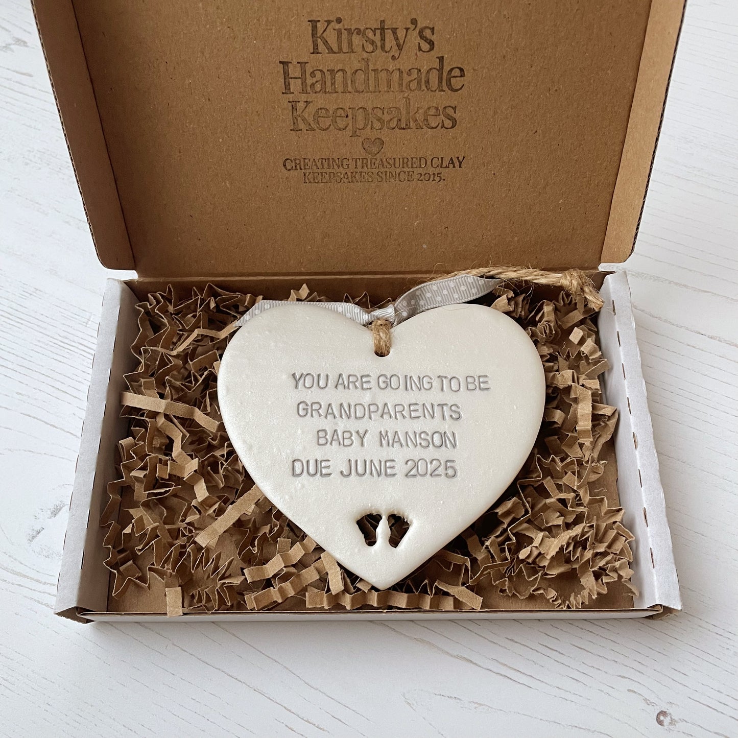 Pearlised white clay hanging heart with baby feet cut out of the bottom and grey personalisation, the heart is personalised with YOU ARE GOING TO BE GRANDPARENTS BABY MANSON DUE JUNE 2025