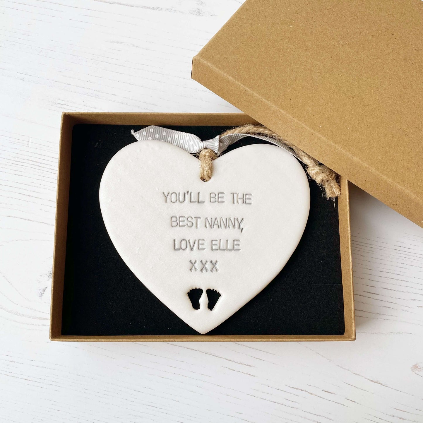 Pearlised white clay hanging heart pregnancy reveal with baby feet cut out of the bottom and grey personalisation, the heart is personalised with YOU’LL BE THE BEST NANNY, LOVE ELLE XXX